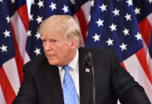 A man in a suit speaking at a podium with American flags in the background