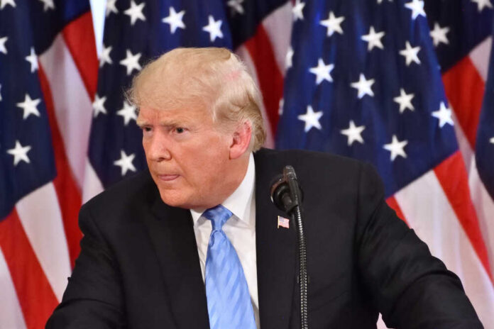 A man in a suit speaking at a podium with American flags in the background