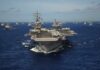 An aircraft carrier surrounded by various naval ships in the ocean