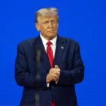 Man in a suit clapping hands on a stage with a blue background
