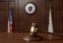 A wooden gavel resting on a table in a courtroom with flags in the background