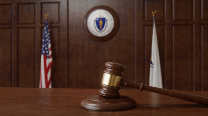 A wooden gavel resting on a table in a courtroom with flags in the background