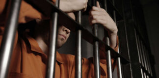 A male prisoner in an orange jumpsuit holding onto the bars of a jail cell