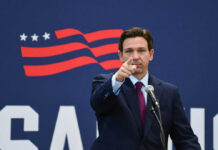 A man in a suit pointing while speaking into a microphone against a backdrop featuring an American flag design