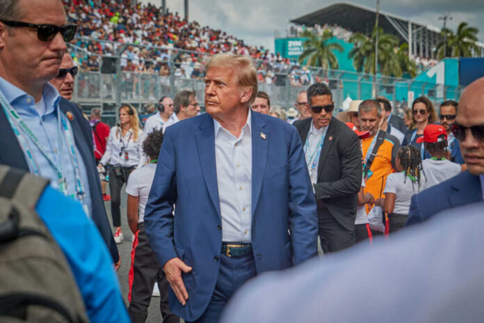 shutterstock_2458373861.jpg A man in a blue suit walking through a crowded outdoor event with security personnel