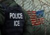 A police officer in a tactical vest stands near a barbed wire fence with an American flag in the background