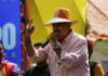 A man in a straw hat passionately speaking into a microphone at a lively outdoor event