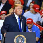 A speaker at a political rally smiling in front of an enthusiastic crowd wearing red hats