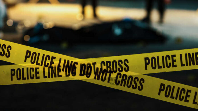 shutterstock_2230640045 Yellow police caution tape at a crime scene with blurred figures in the background