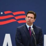 A man in a suit standing at a microphone with a serious expression against a backdrop featuring a stylized American flag