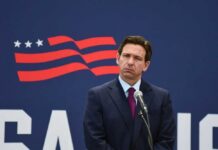 Property Taxes Axed? DeSantis Drops Bomb A man in a suit standing at a microphone with a serious expression against a backdrop featuring a stylized American flag
