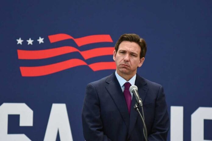 Rochester,,N.h.usa,-,July,31,,2023:,Florida,Gov.,Ron,Desantis A man in a suit standing at a microphone with a serious expression against a backdrop featuring a stylized American flag