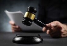 A gavel held above a sounding block with a person reading documents in the background