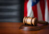 Court Crackdown Exposes Biden Recordings A wooden gavel resting on a round base with a blurred American flag in the background