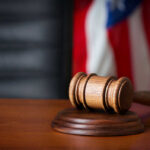 A wooden gavel resting on a round base with a blurred American flag in the background
