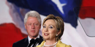 Hillary Clinton speaking at a podium with Bill Clinton in the background