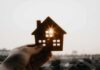Federal Squeeze Hits Anti-Housing Rules Hand holding a small house cutout against a bright sunset sky