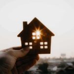 Federal Squeeze Hits Anti-Housing Rules Hand holding a small house cutout against a bright sunset sky