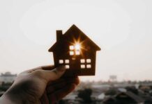 Federal Squeeze Hits Anti-Housing Rules Hand holding a small house cutout against a bright sunset sky