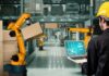 Worker with laptop overseeing robotic arms moving boxes in warehouse