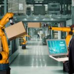 Worker with laptop overseeing robotic arms moving boxes in warehouse
