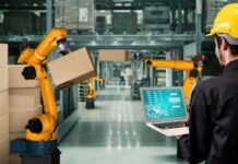 AI Arms Factory Jackpot Raises Eyebrows Worker with laptop overseeing robotic arms moving boxes in warehouse