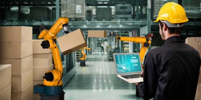 shutterstock_2025507059 Worker with laptop overseeing robotic arms moving boxes in warehouse
