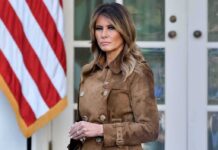 Melania Trump standing in a brown suede coat near an American flag