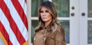 Melania Trump standing in a brown suede coat near an American flag