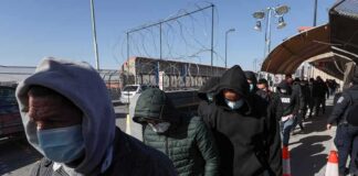 People in hoodies and masks walk in a line along a fenced walkway