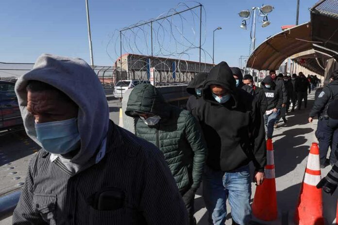 shutterstock_2089375453 People in hoodies and masks walk in a line along a fenced walkway